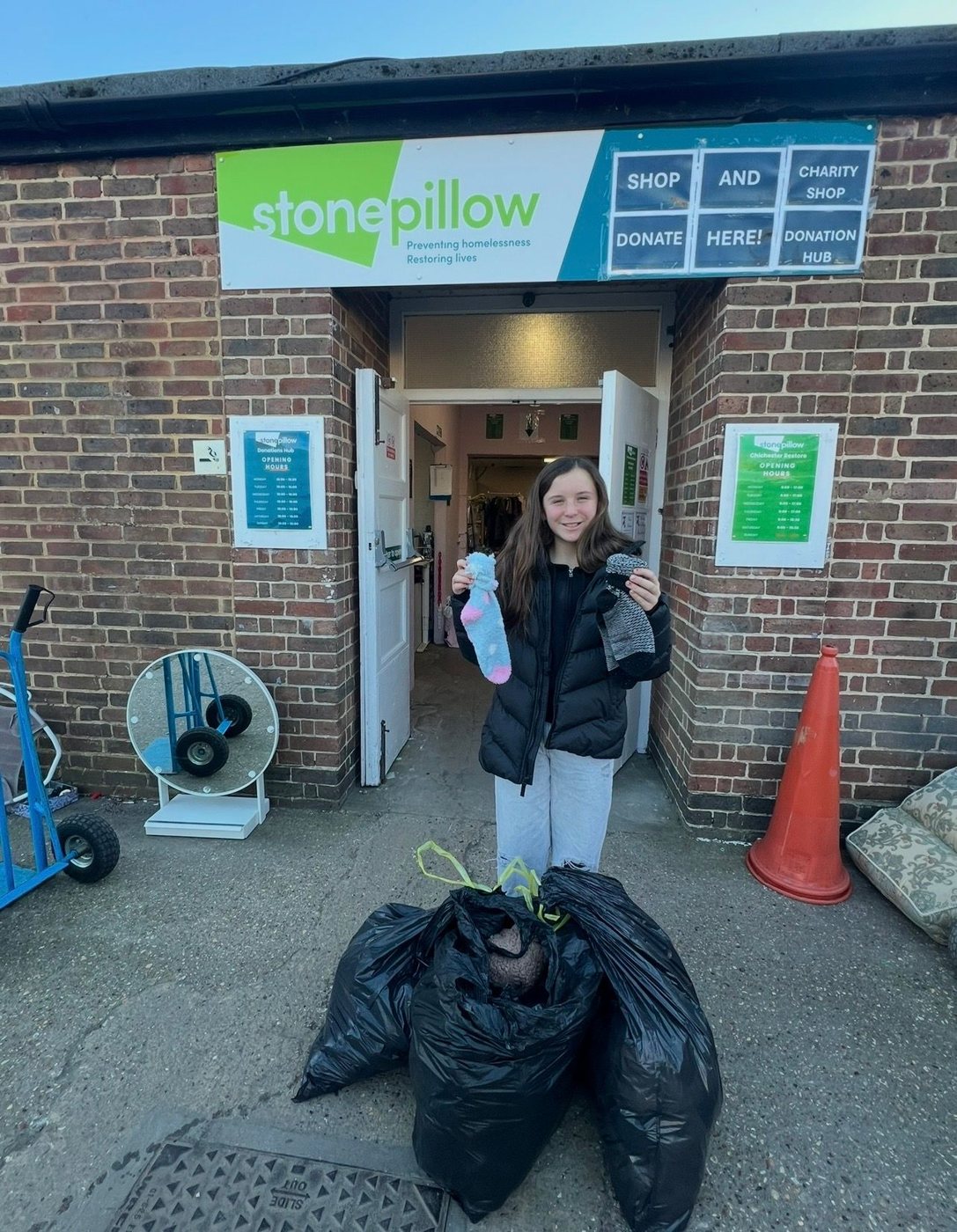 Elsie outside the Stonepillow donations hub holding up two socks, with full bags of socks at her feet